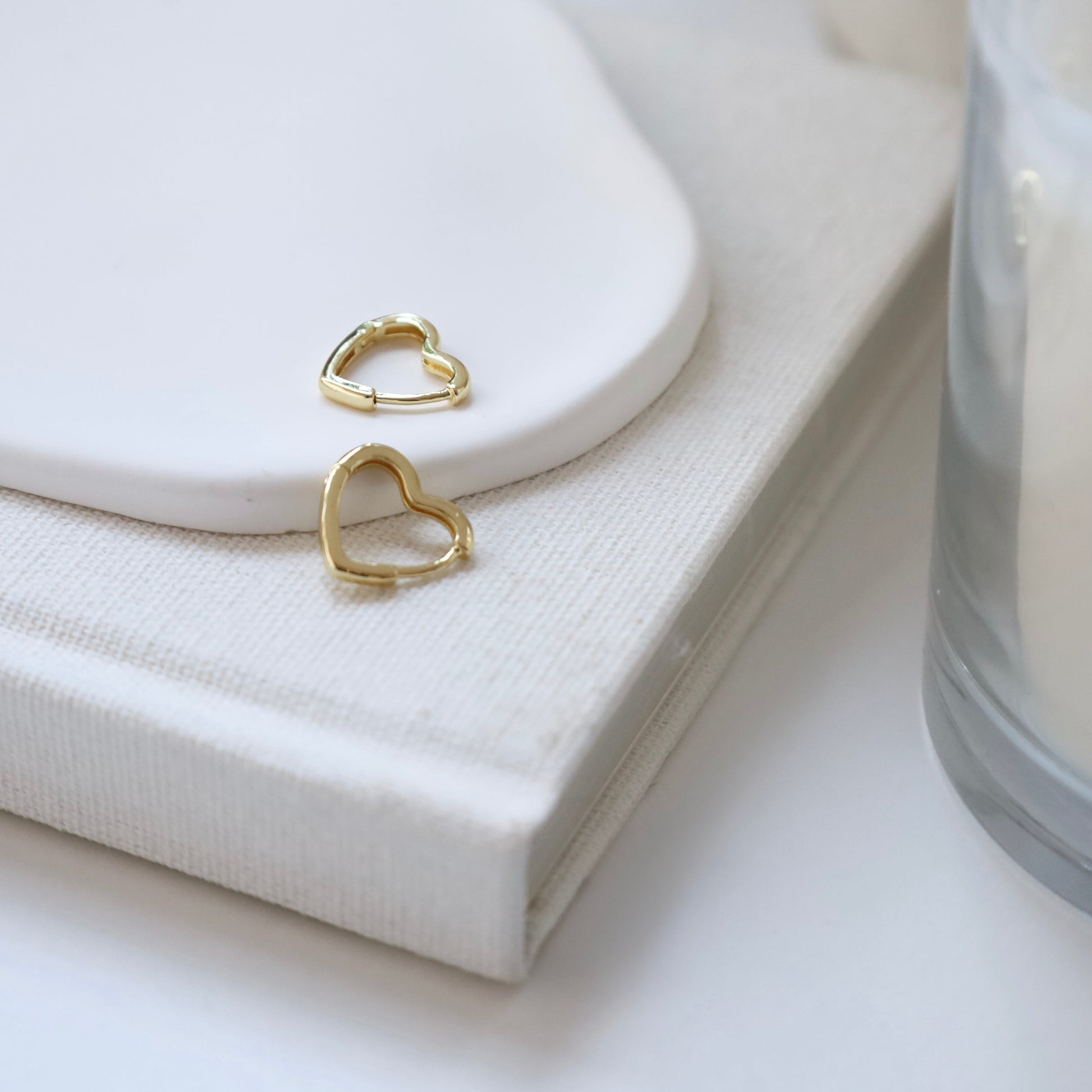 Gold heart-shaped earrings on a white surface with a candle and a book in the background.