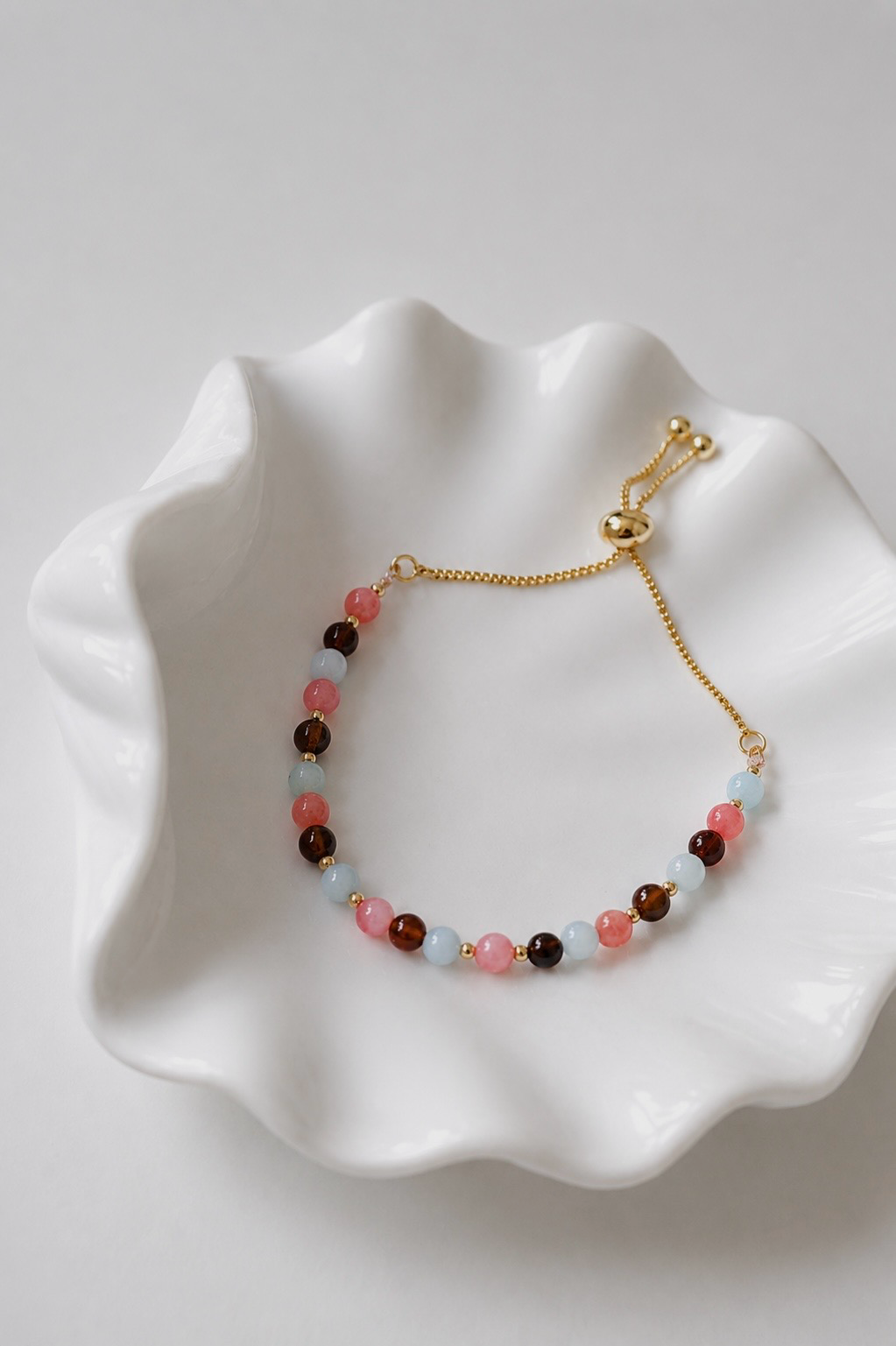 Gold adjustable mixed gemstone bracelet with pink coral, aquamarine and brown agate gemstones displayed in a white ceramic shell dish. Elegant everyday jewellery from the SophieJean Summer Essentials collection.
