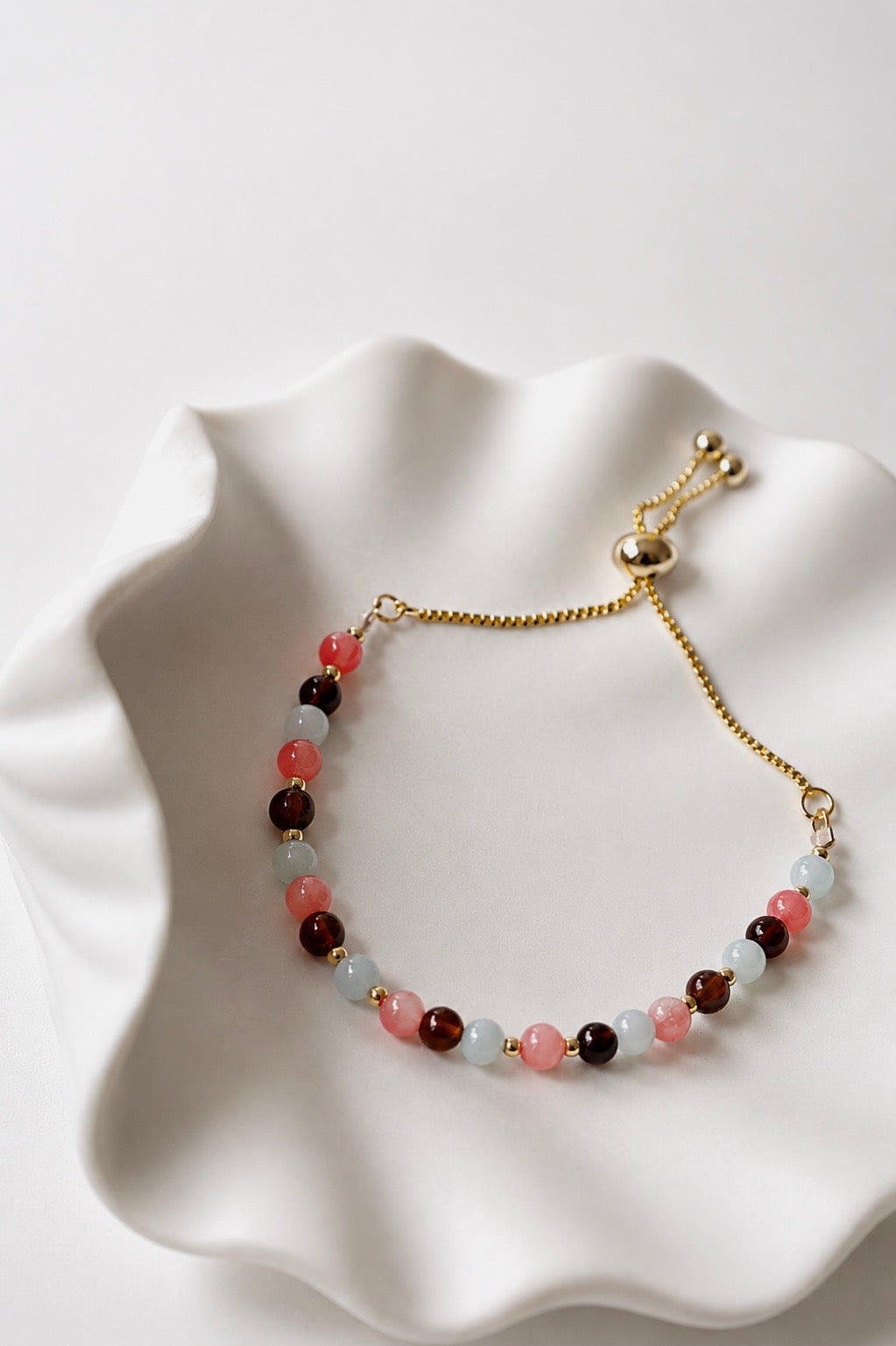 Gold adjustable mixed gemstone bracelet with pink coral, aquamarine and brown agate gemstones displayed in a white ceramic shell dish. Coastal-inspired everyday jewellery from the SophieJean Summer Essentials collection