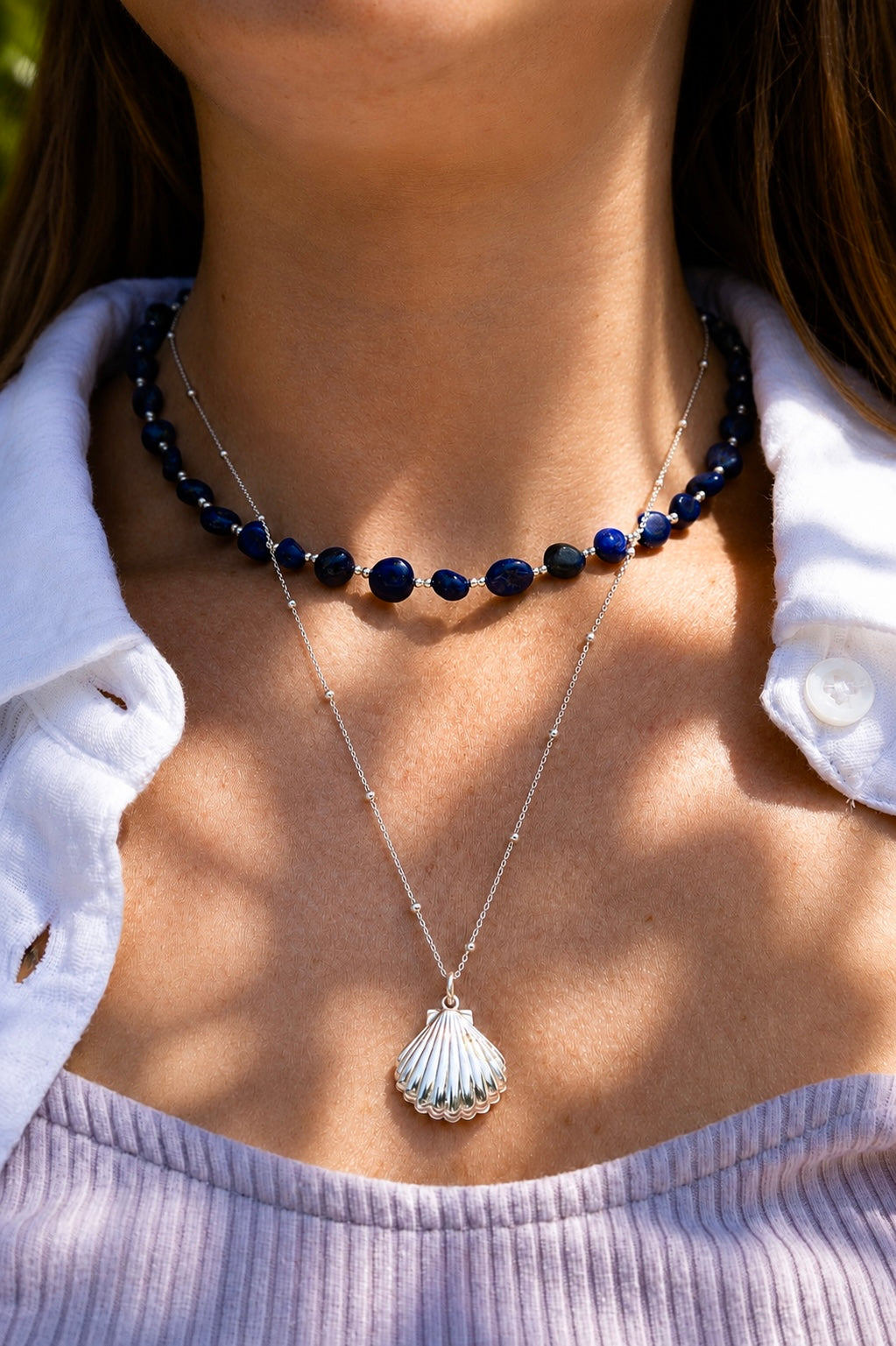 A close-up of a person wearing layered necklaces: a short beaded necklace with dark blue beads and a longer silver chain necklace with a scallop shell locket pendant, styled with a white collared shirt.