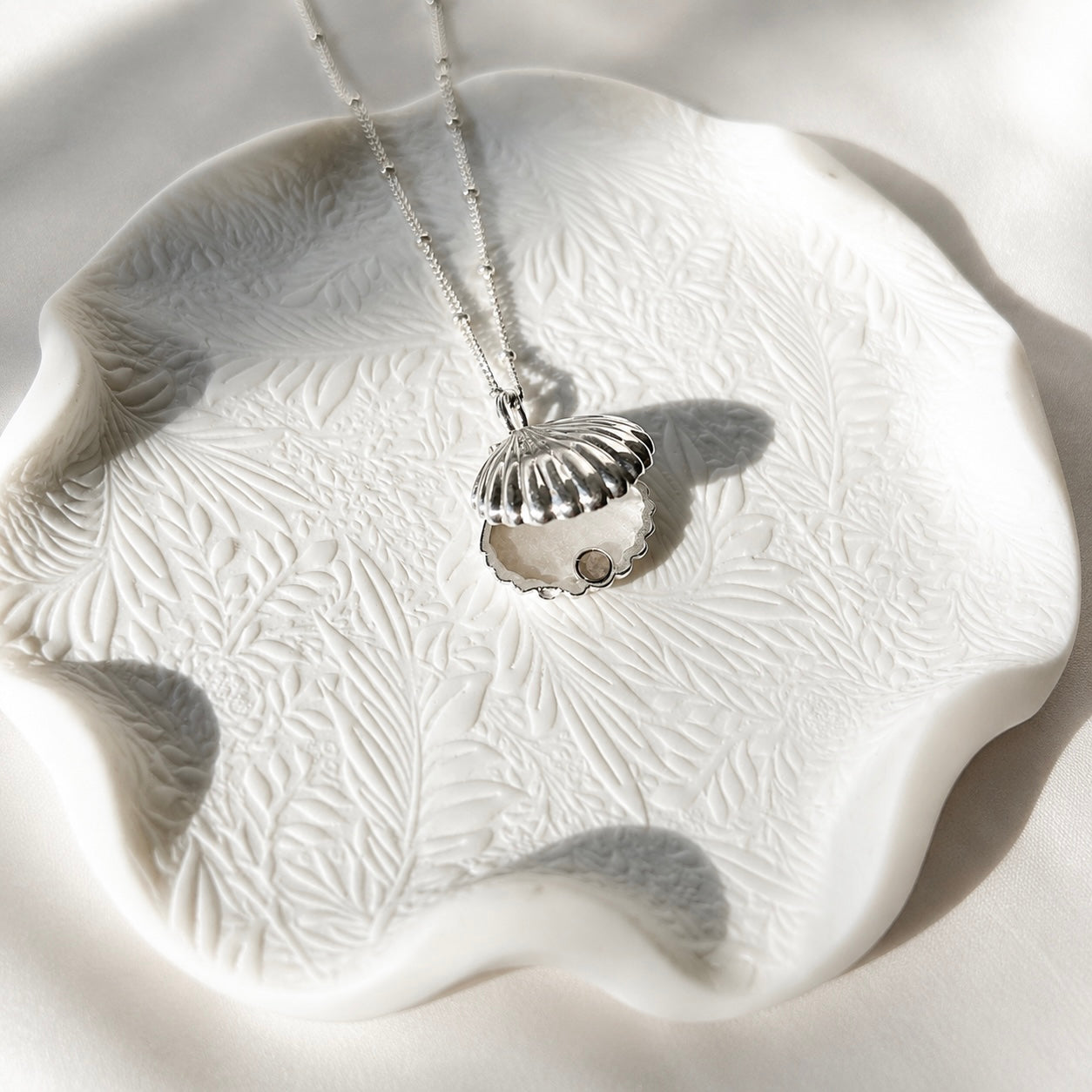 A silver shell-shaped opened locket necklace with a beaded chain, displayed on a white textured ceramic dish with leaf patterns under bright sunlight.